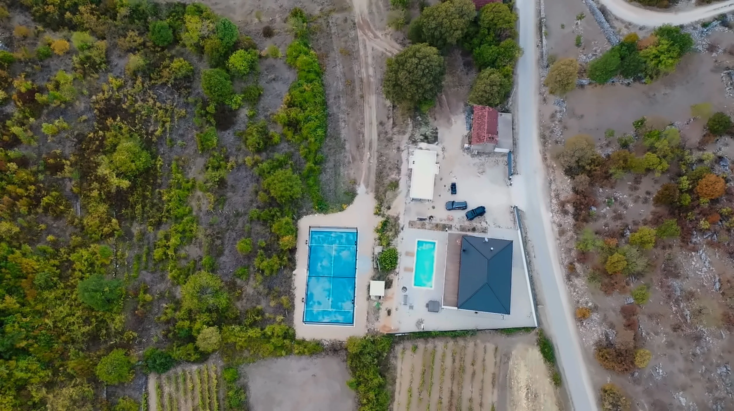 Pogled odozgo na Padel Hercegovina u Drinovcima: plavi padel teren, bazen, kuća i parking uz cestu, okruženi vinogradima i šumom